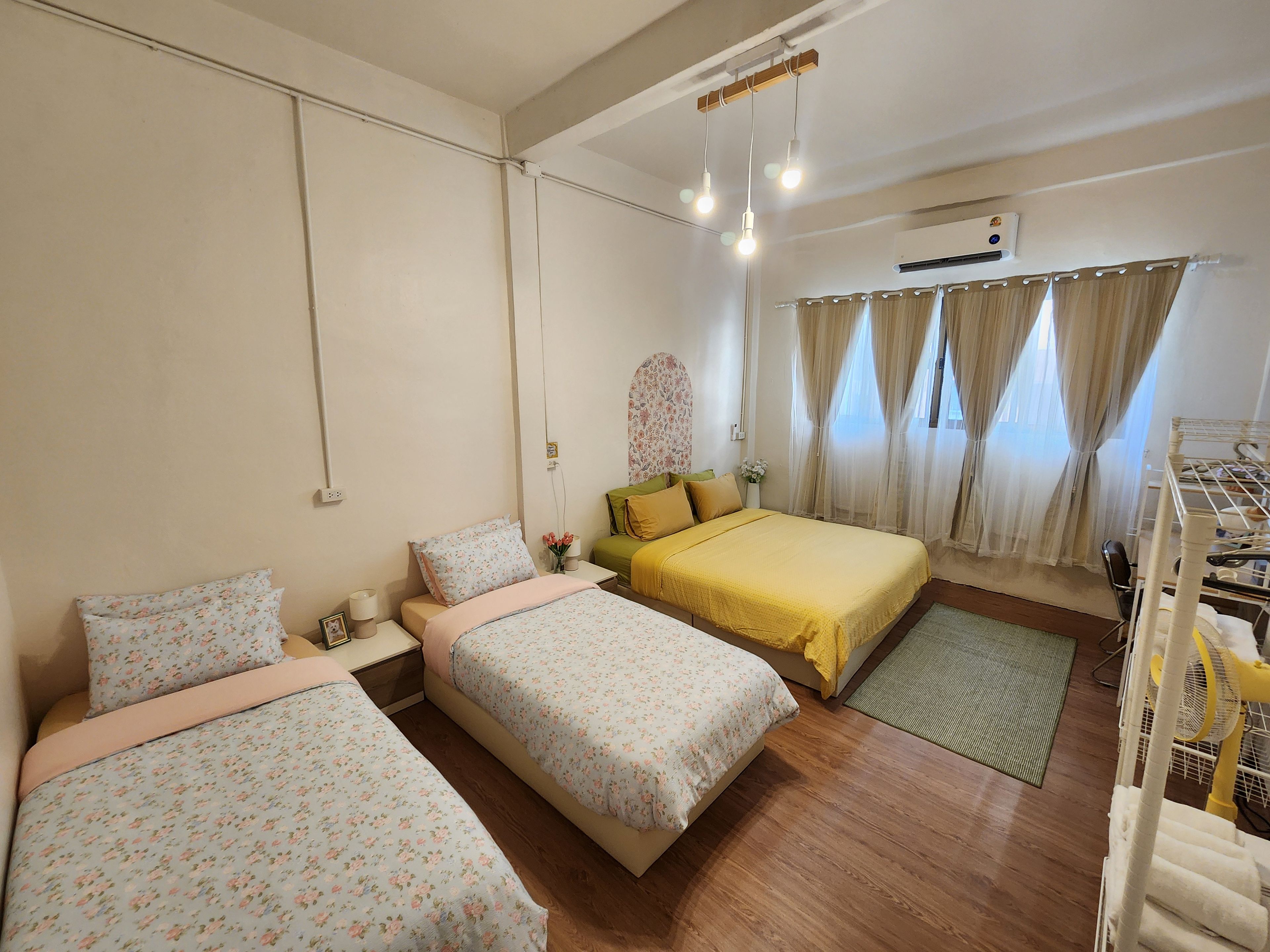 A room with two beds, a bedside table, a lamp, a window with curtains, and an air conditioning unit.