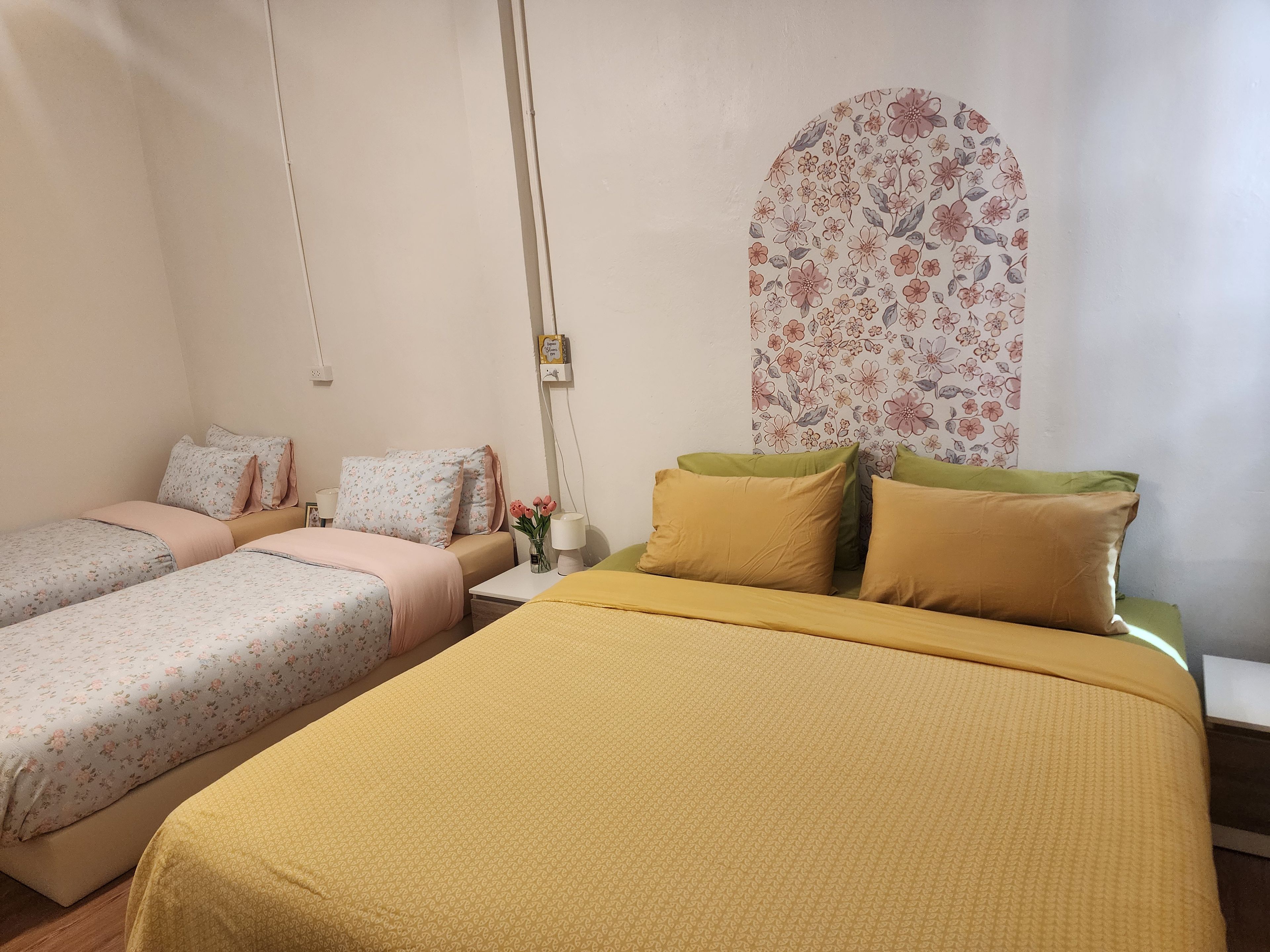 A hotel room with two single beds, a large bed, a nightstand, a lamp, and a floral-patterned wall.