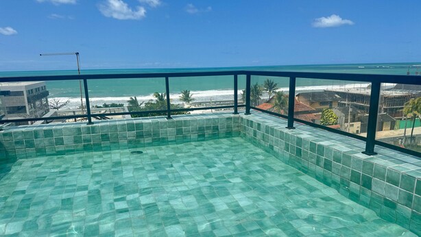 Cielo azul despejado, piscina con piso de azulejos y vista al océano y edificios al fondo.