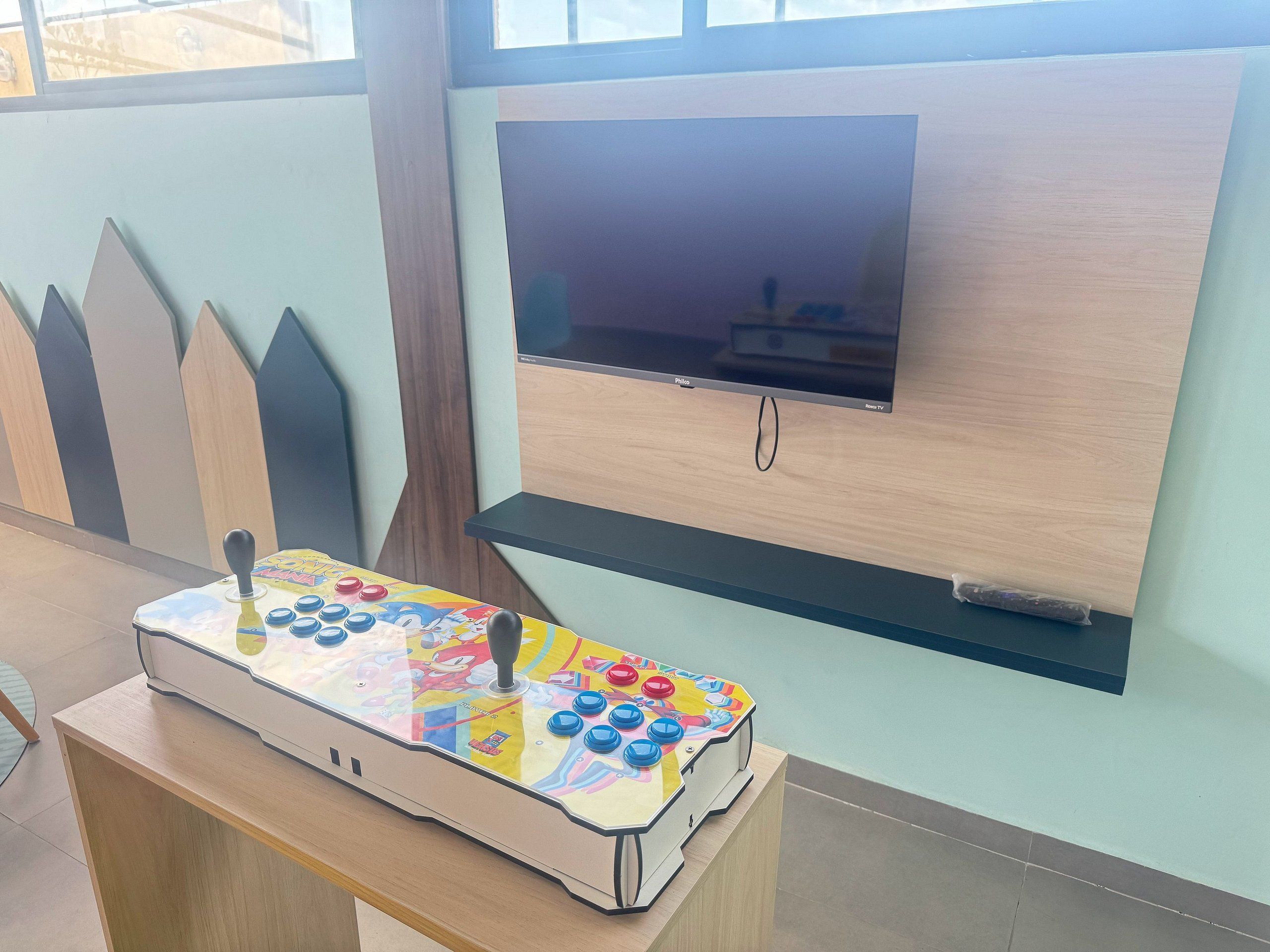 Une pièce avec un téléviseur fixé au mur, une table sur laquelle se trouve un jeu d’arcade coloré, et un meuble en bois.