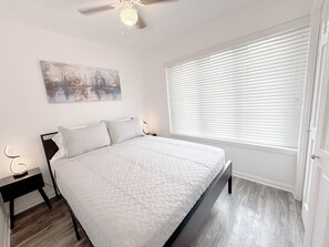 Een slaapkamer met een bed, een plafondventilator, een nachtkastje met een lamp, een raam met zonwering en een aan de muur hangend schilderij.