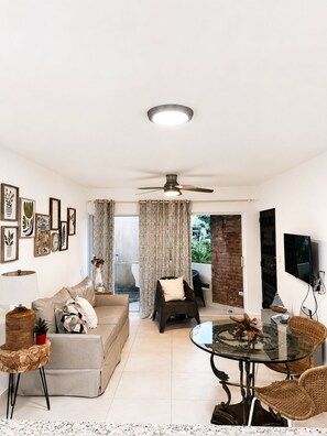 Ein Wohnzimmer mit einer Couch, einem Glastisch, Korbstühlen, einem Deckenventilator und einem an der Wand montierten Fernseher.
