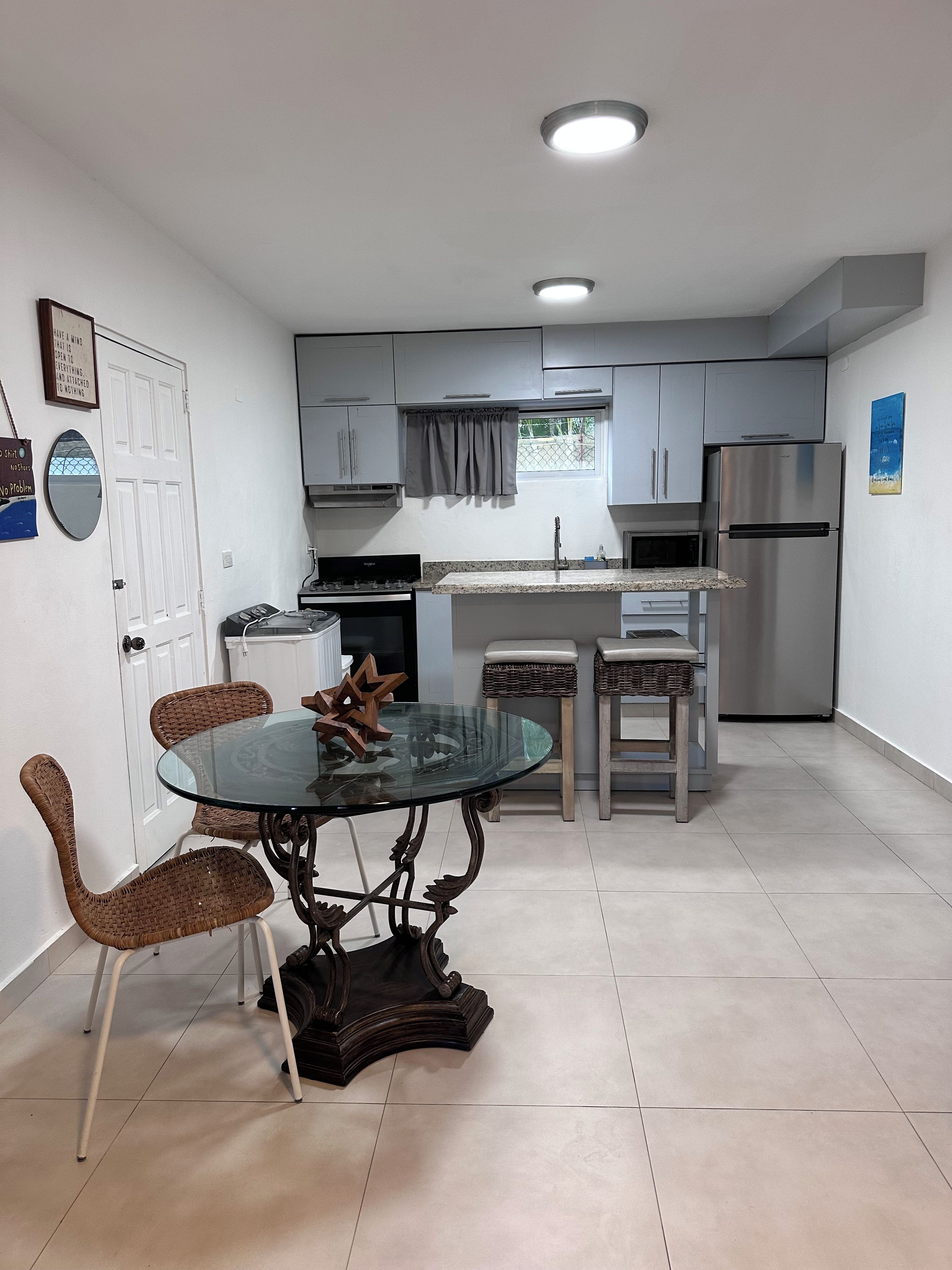 Una cocina compacta con muebles blancos, un refrigerador de acero inoxidable, una mesa de comedor con cubierta de vidrio y sillas de mimbre, y una pequeña ventana con cortina.