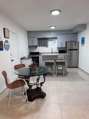A compact kitchen with white cabinets, a stainless steel refrigerator, a glass-top dining table with wicker chairs, and a small window with a curtain. - Cozy 1-bedroom apartment in beautiful Costambar with AC (Costambar)