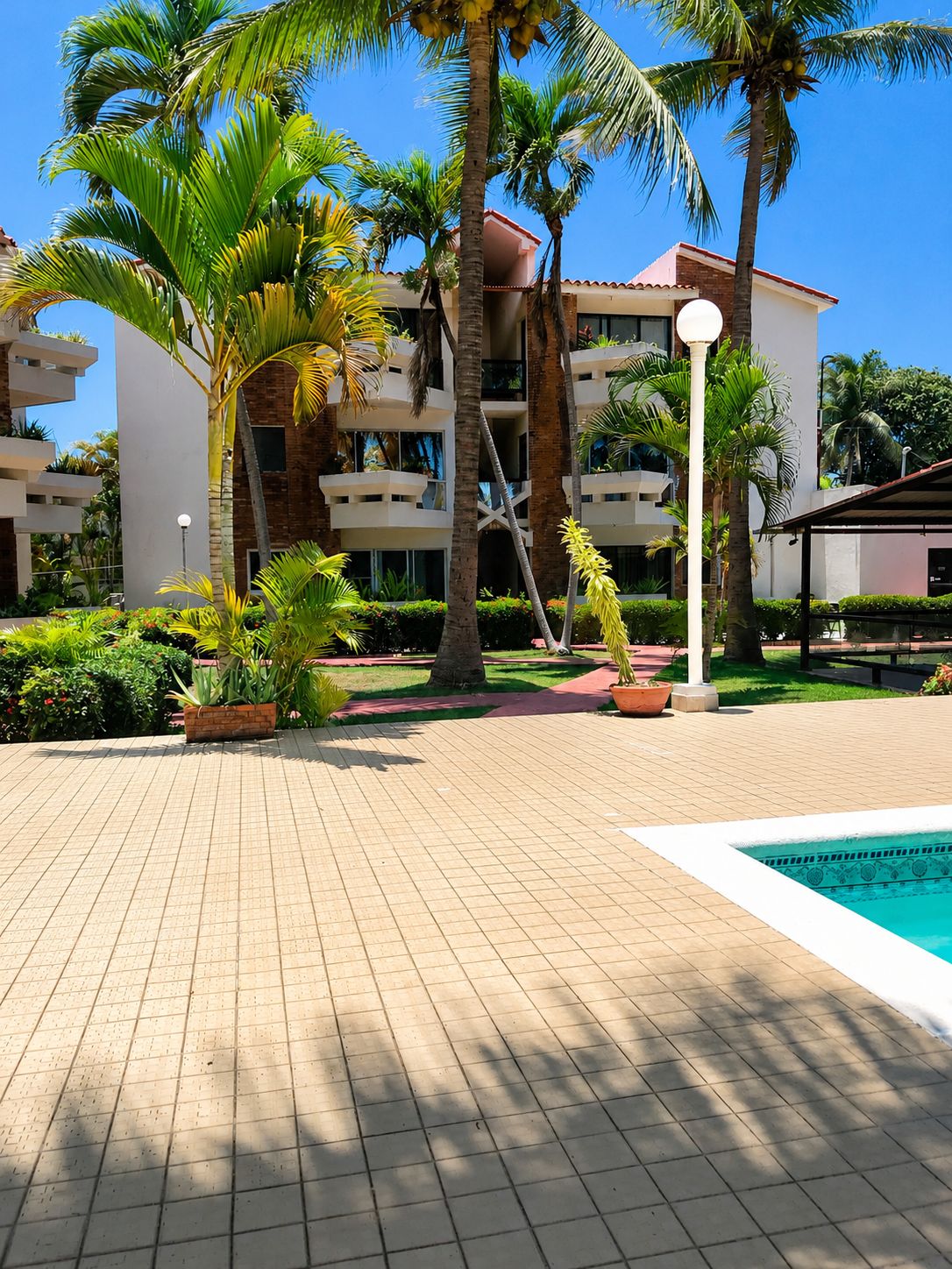 Une cour d’hôtel avec un sol carrelé, des palmiers, une piscine et un immeuble à plusieurs étages doté de balcons.