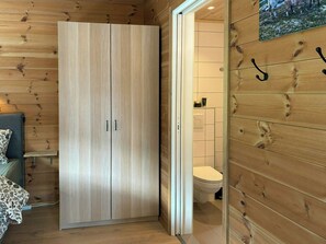 Une pièce aux murs lambrissés, comprenant une armoire, un lit recouvert d’une couvre-lit à motifs floraux et des toilettes.