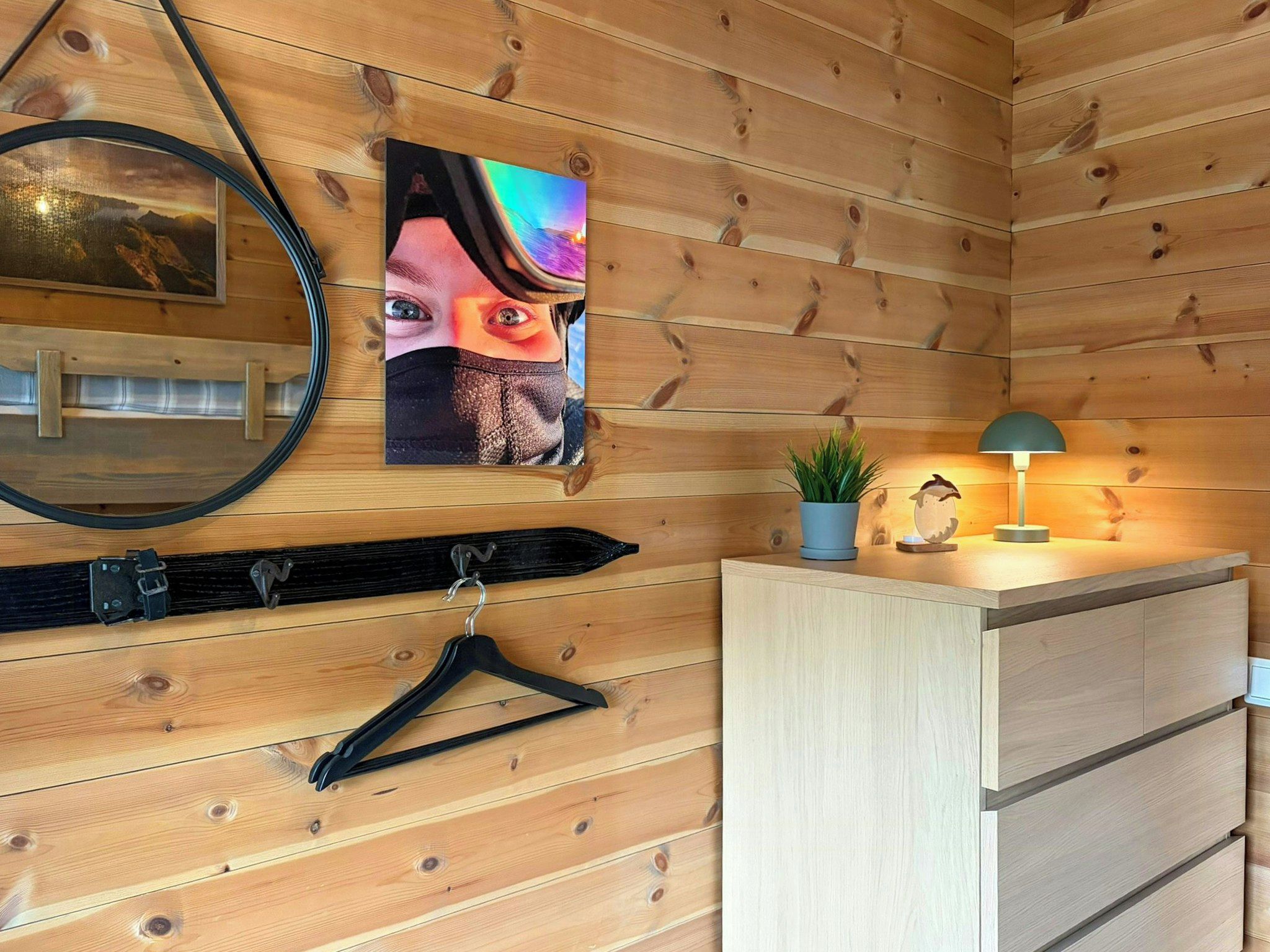 A wooden cabin interior with a mirror, a framed picture, a hanging coat rack, a small plant, and a desk with a lamp.