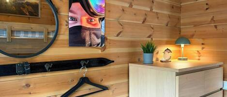 A wooden cabin interior with a mirror, a framed picture, a hanging coat rack, a small plant, and a desk with a lamp.