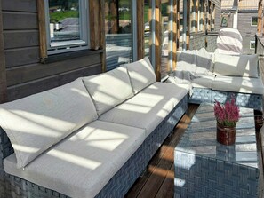 A long, light-colored sofa with a woven base, positioned on a wooden deck with a patterned tile floor.