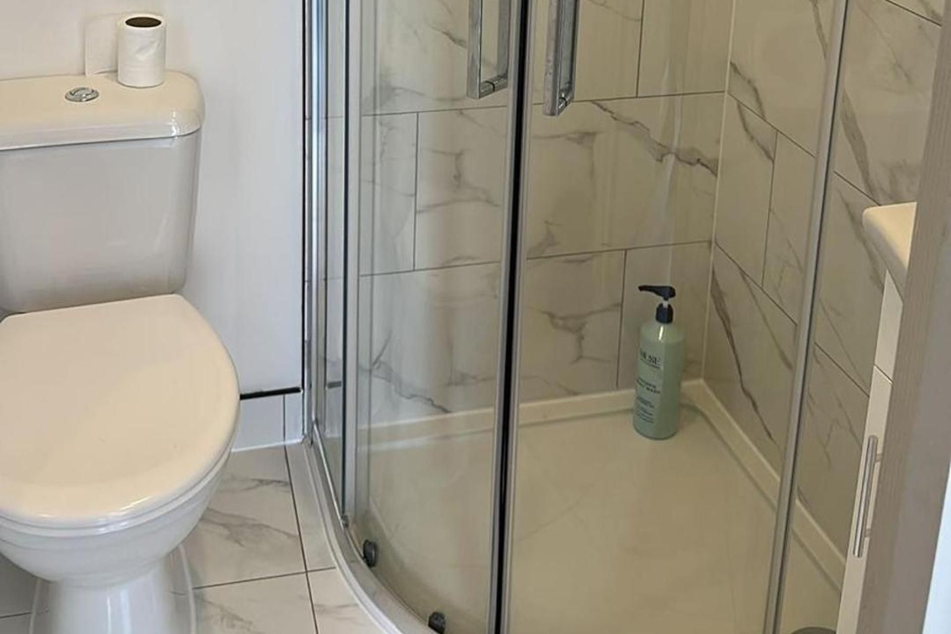 A bathroom with a toilet, a glass shower enclosure, and a bottle of shampoo on the floor.