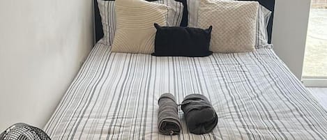 Cama com roupa de cama listrada e várias almofadas, janela com cortinas e vista para uma cerca de madeira lá fora.