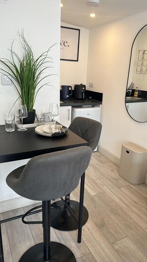 A modern dining area with a table set for two, a potted plant, and a kitchen in the background. - Studio Sleeps 2 With Wifi (Thornton Heath)