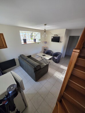 A modern living room with a sofa, dining table, and chairs, a TV mounted on the wall, and a staircase leading to another level.