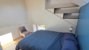 A modern bedroom with a bed, a chair, a bedside table with a lamp, and a window. - Cozy Home (Bonnemain)