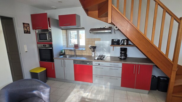 A modern kitchen with red cabinets, a wooden staircase, and a dining area with a table and chairs. - Cozy Home (Bonnemain)