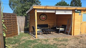 A wooden outdoor seating area with a table and chairs, a sign reading 'LA FORGE,' and a wooden fence in the background. - Cozy Home (Bonnemain)