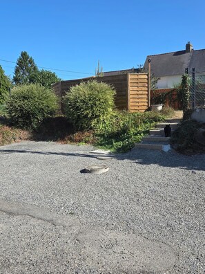 A gravel driveway leading to a wooden gate, surrounded by dense shrubbery and a clear blue sky.