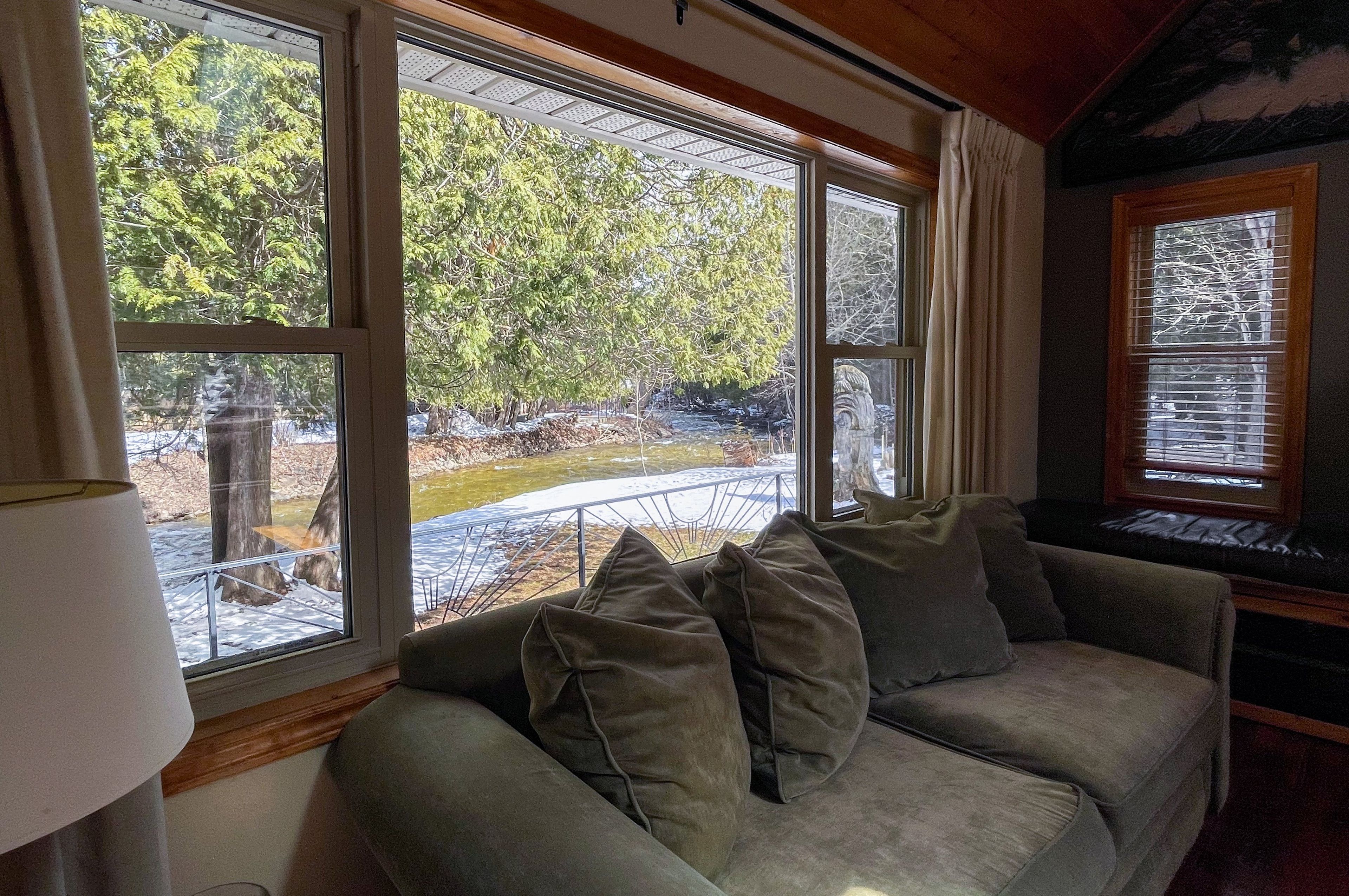 A room with a sofa, a lamp, and large windows offering a view of trees and a river.