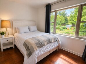 Ein Schlafzimmer mit einem Bett, einem Nachttisch, einer Lampe und einem Fenster mit Blick auf Grünflächen.