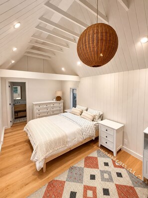 Ein Schlafzimmer mit einem großen Bett, weißen Nachttischen, einer gewobenen Pendelleuchte und einem gemusterten Teppich.