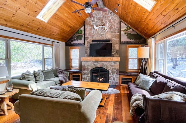 A cozy cabin interior with a stone fireplace, wooden ceiling, and large windows.