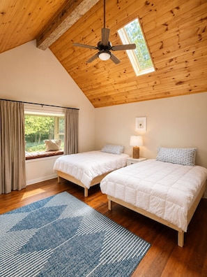 A bedroom with two beds, a ceiling fan, a window with curtains, and a view of greenery outside.