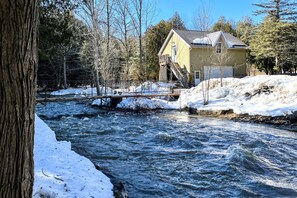 Ein Haus an einem Fluss mit Schnee an den Ufern und einer Holzbrücke über dem Wasser.