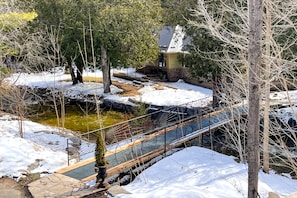 A snow-covered outdoor area with a wooden bridge, a stone pathway, and a small pond.