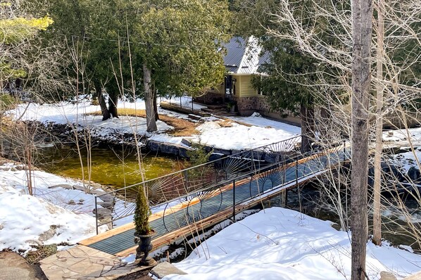 A snow-covered outdoor area with a wooden bridge, a stone pathway, and a small pond.