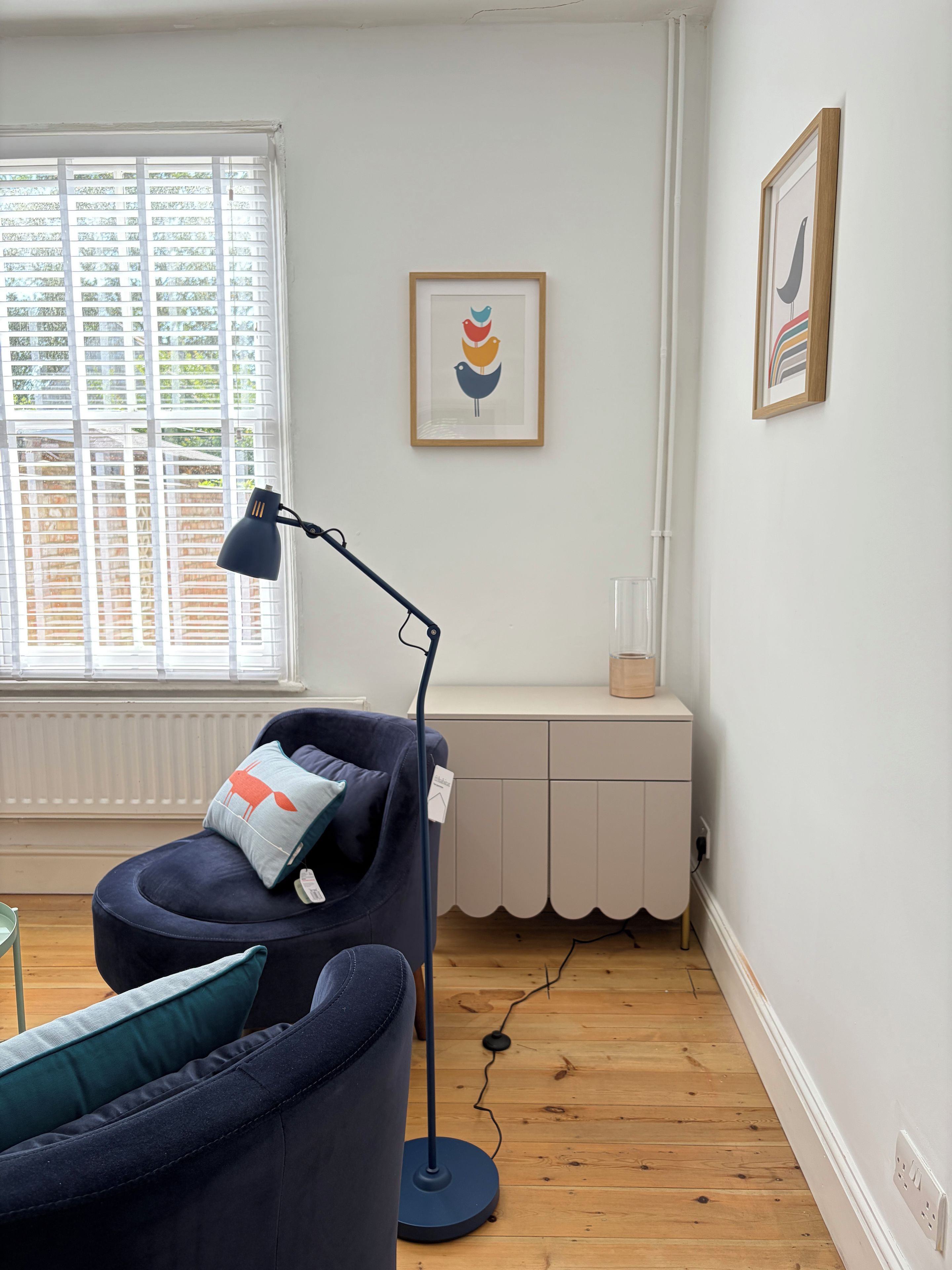 A room with a blue chair, a floor lamp, a radiator, a window with blinds, and two framed pictures on the wall.