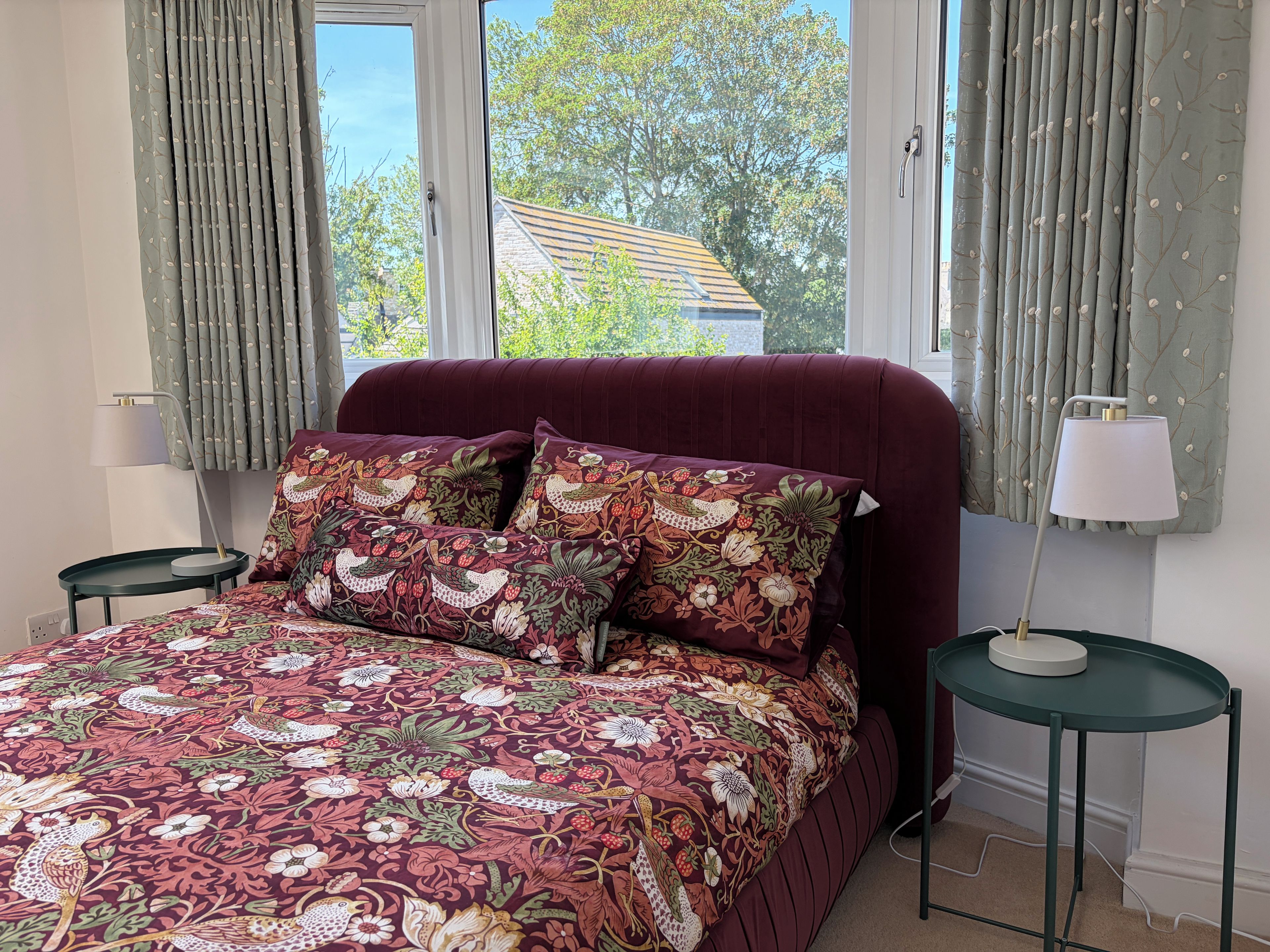 A bedroom with a bed, two nightstands, a window with curtains, and a view of a house outside.