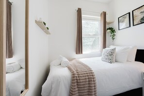 Un lit bien fait, avec du linge de lit blanc et une couverture marron à rayures, une plante sur la table de chevet et une fenêtre avec des rideaux.