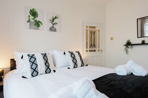 A hotel room with a bed, pillows, a nightstand, a lamp, a door, and wall art. - 4-Bed Home | 7 Beds | Free Parking | Groups Welcome (Manchester)