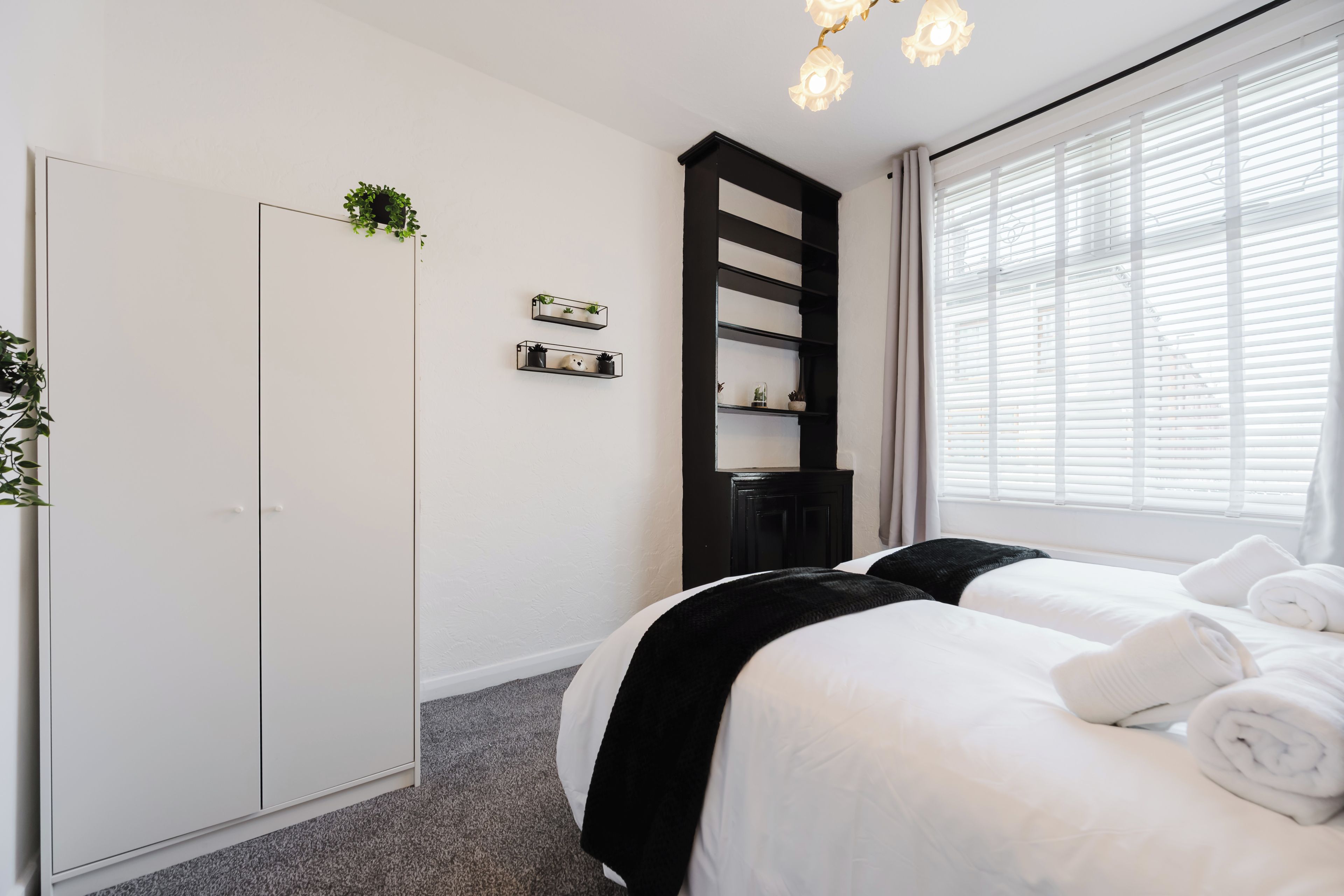 A hotel room with a bed, a wardrobe, a shelf with plants, and a window with blinds.
