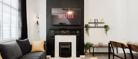 A modern living room with a fireplace, a sofa, a dining table, and a television mounted above the fireplace displaying 'NETFLIX'.
