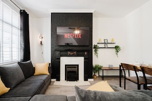 A modern living room with a fireplace, a sofa, a dining table, and a television mounted above the fireplace displaying 'NETFLIX'.