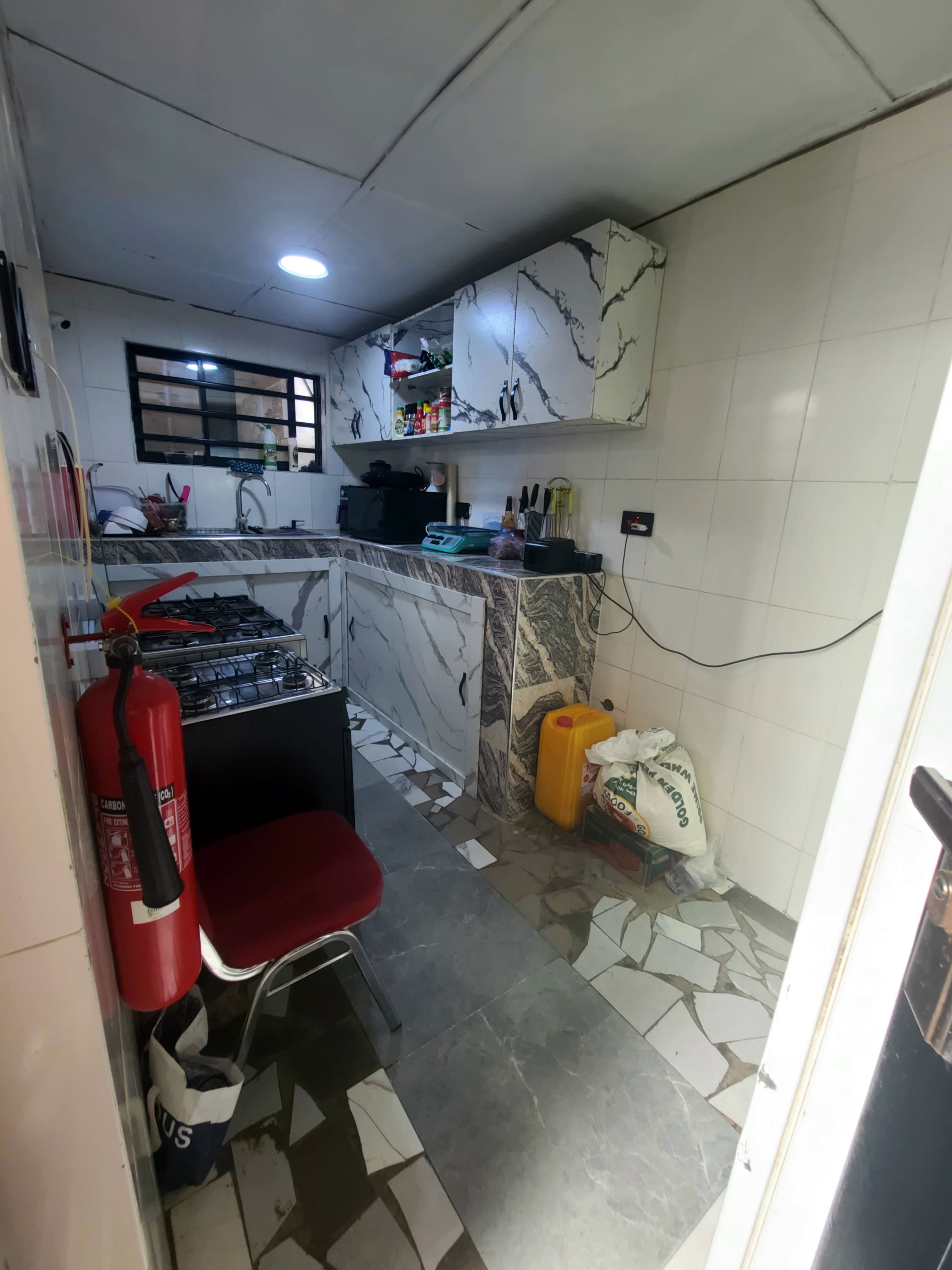A compact kitchen with a gas stove, a red chair, a fire extinguisher, and a yellow container.