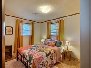 Quarto com cama, duas luminárias de cabeceira, janela com cortinas, um quadro na parede e uma pequena prateleira.