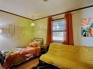 A bedroom with two beds, a sofa, a hanging macrame decoration, a lamp, and a window with curtains.