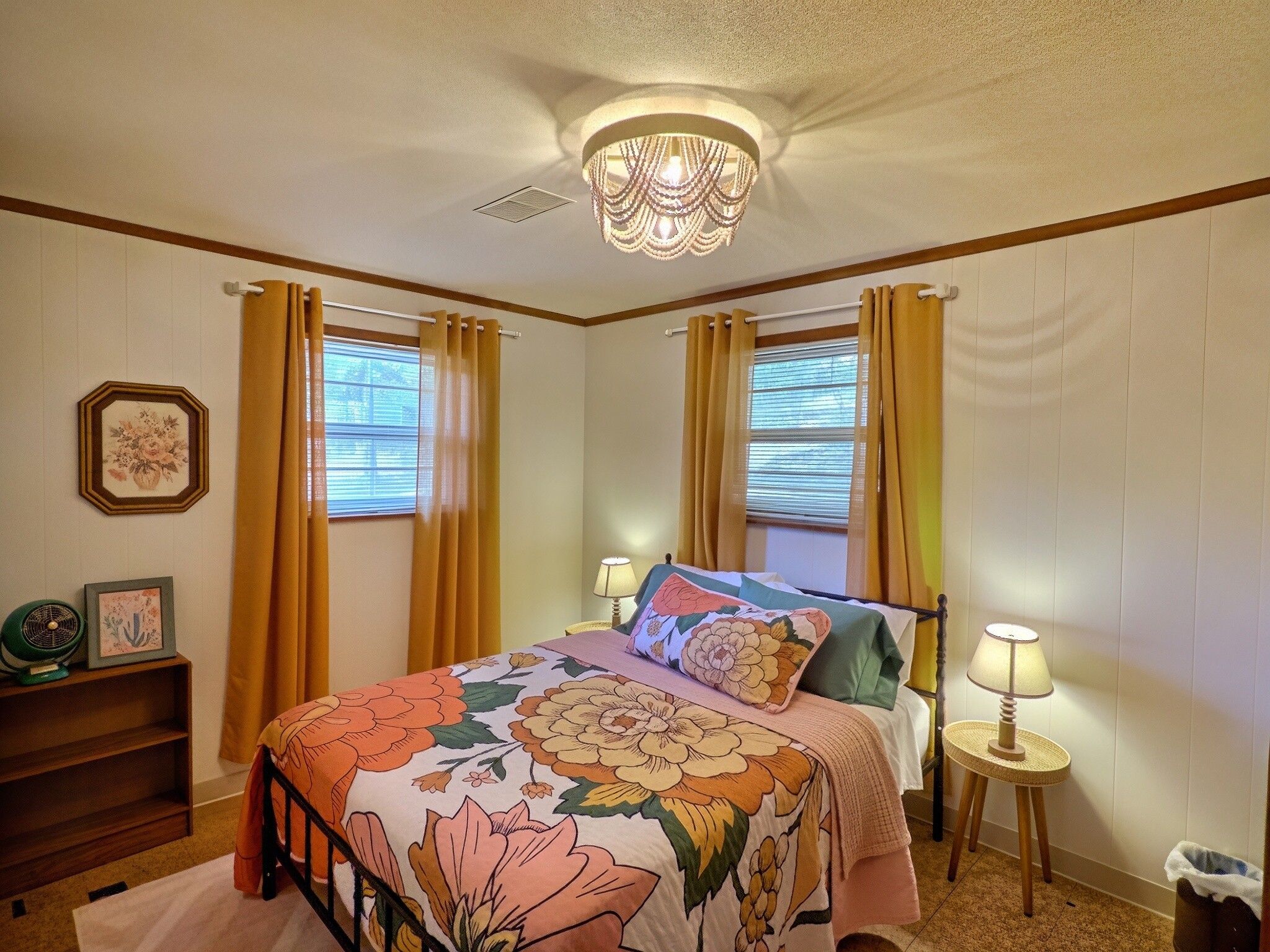 Une chambre à coucher avec une couvre-lit à motifs floraux, deux lampes de chevet, une table de chevet, un ventilateur et un cadre photo posé sur une étagère.