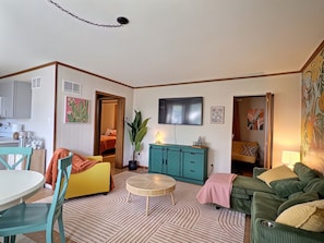 A living room with a television, sofa, chairs, a dining table, and a kitchen area.