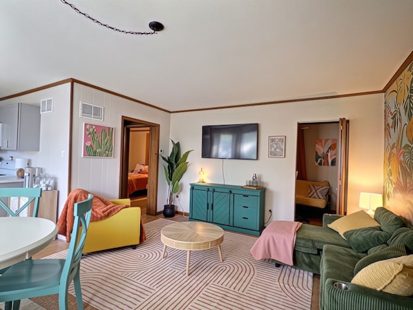 A living room with a television, sofa, chairs, a dining table, and a kitchen area.