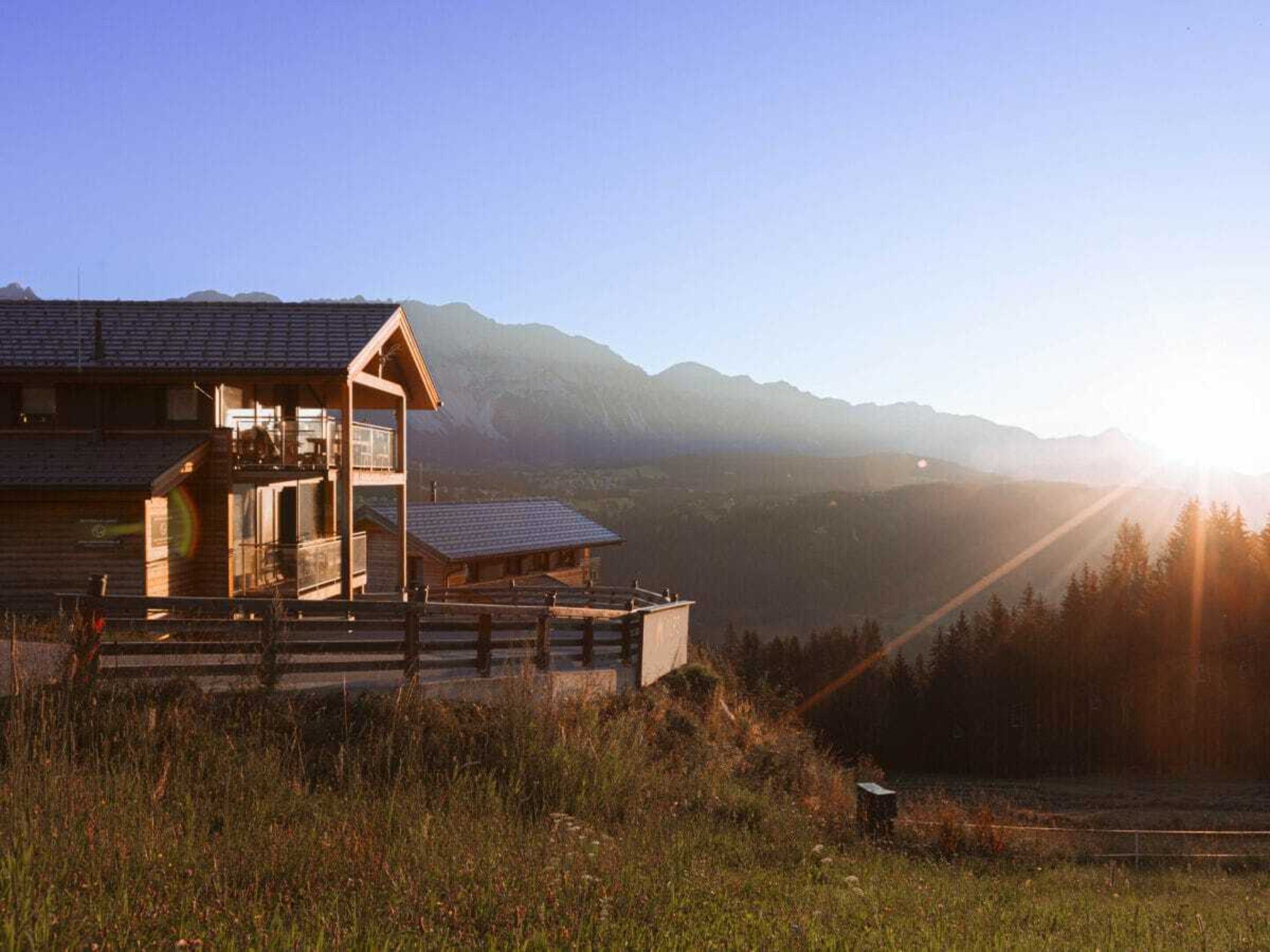 A wooden cabin with a balcony, set against a mountainous backdrop during sunset.