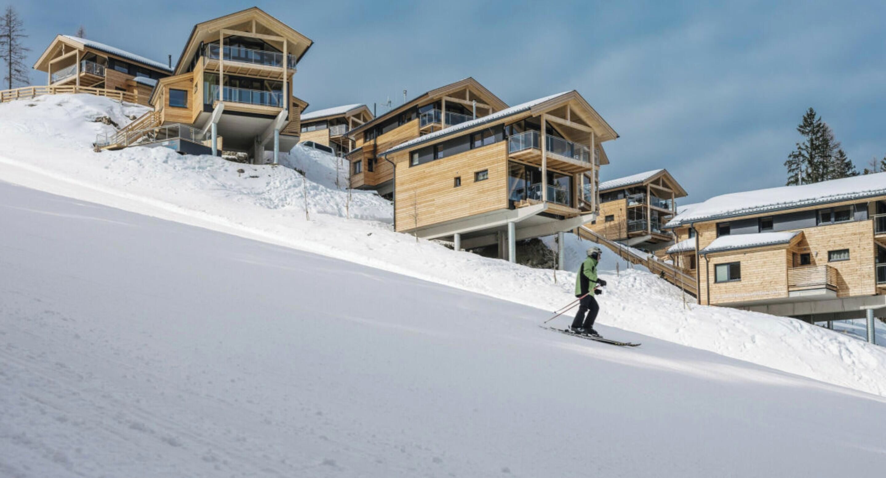 A ski resort with wooden chalet-style buildings, a person skiing down a slope, and a clear sky.