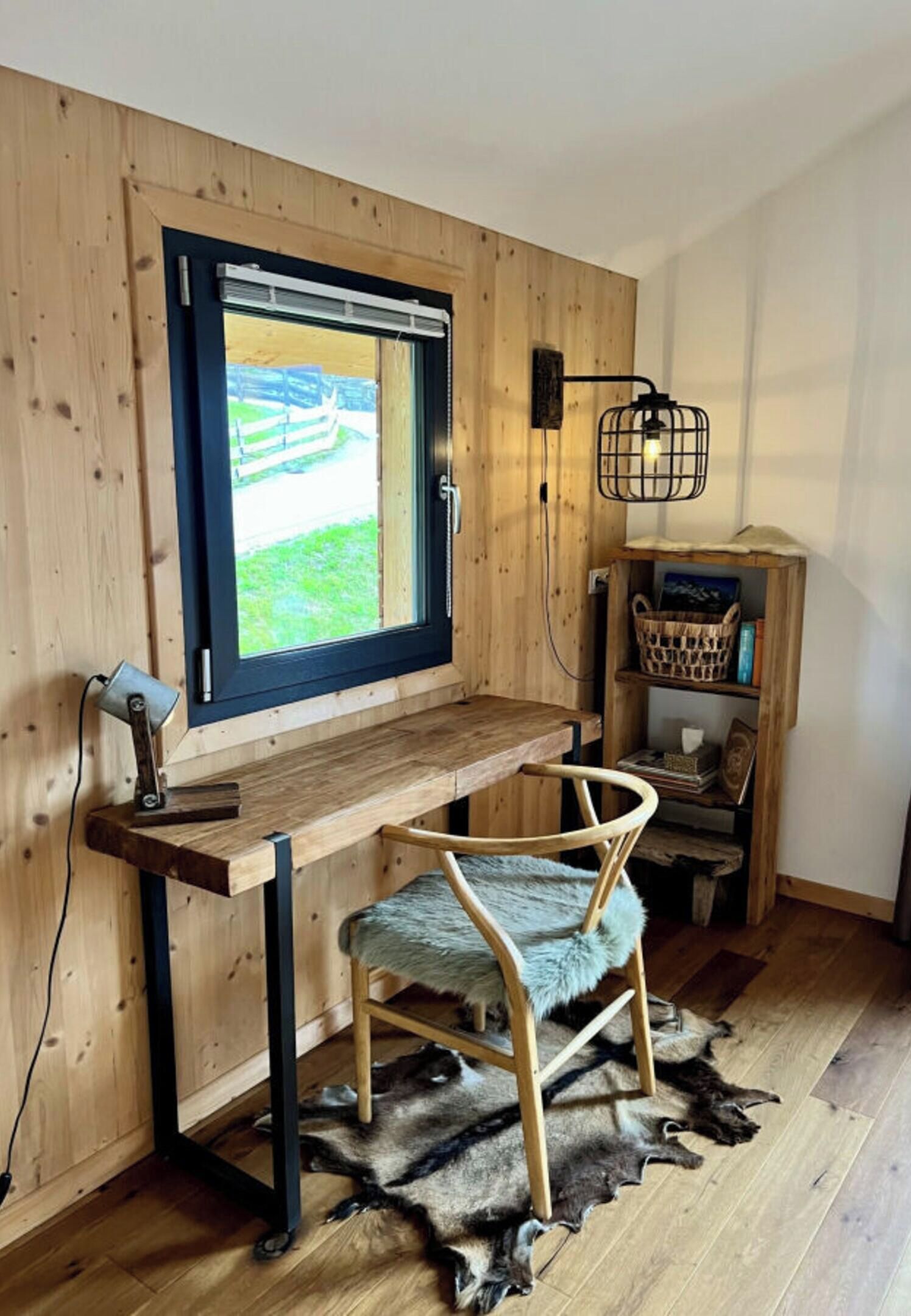 Un bureau en bois, avec une fenêtre, une chaise, une lampe et une étagère remplie de livres et d’un panier.