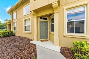 A two-story residential building with a front entrance, a doormat, and a small garden area with mulch and shrubs. - Spacious 3 BR with Pool - Near St. Pete Beaches, Downtown & Tampa (Pinellas Park)