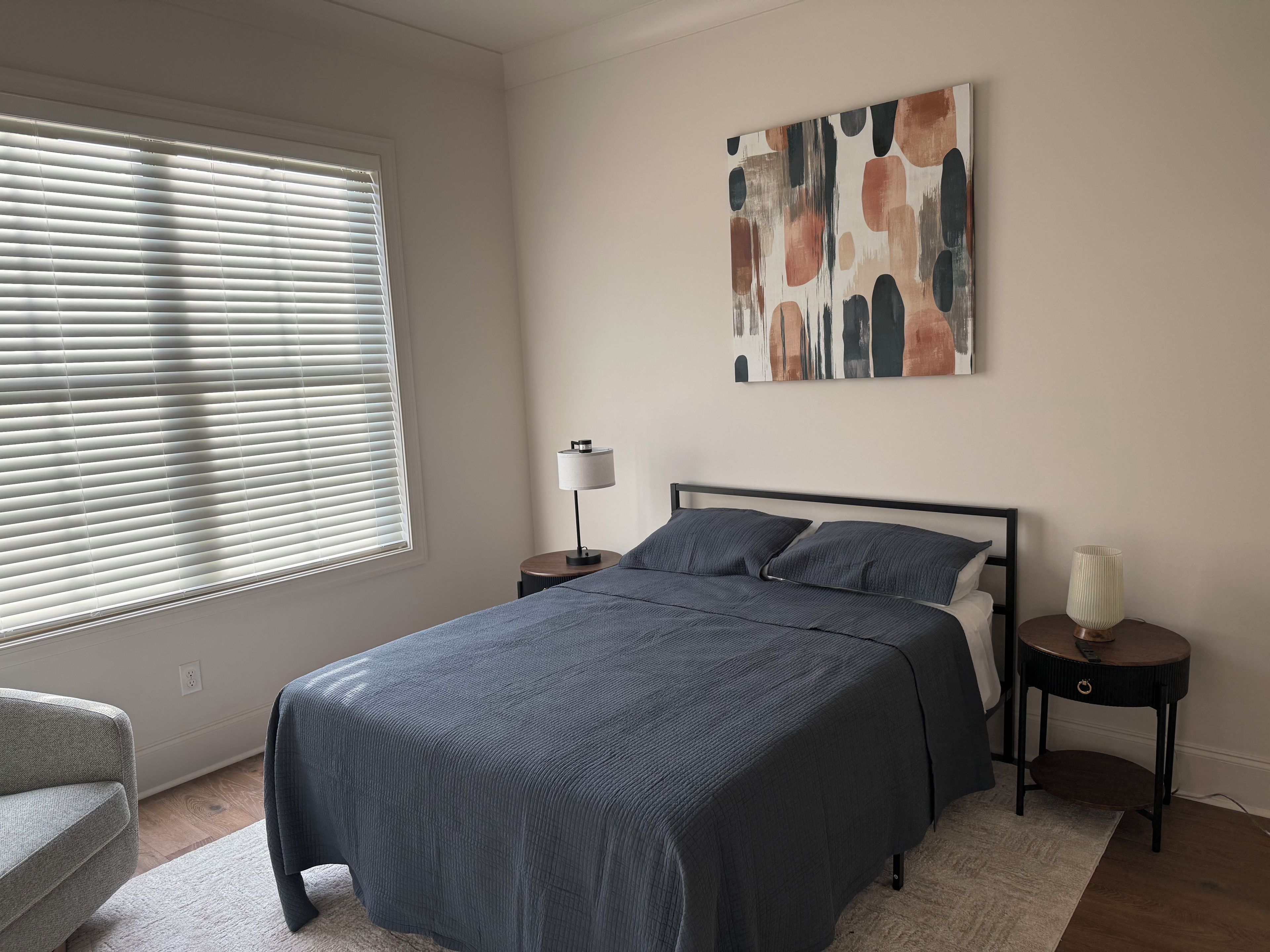 Quarto com cama, mesa de cabeceira, luminária, cadeira e janela com persianas.