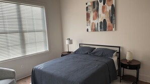 A bedroom with a bed, a bedside table, a lamp, a chair, and a window with blinds. - The Renaissance House, a newly built custom home (Tuskegee)