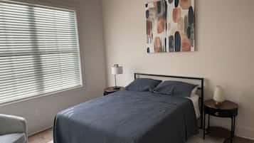 Quarto com cama, mesa de cabeceira, luminária, cadeira e janela com persianas.