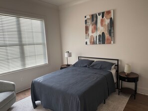 A bedroom with a bed, a bedside table, a lamp, a chair, and a window with blinds.